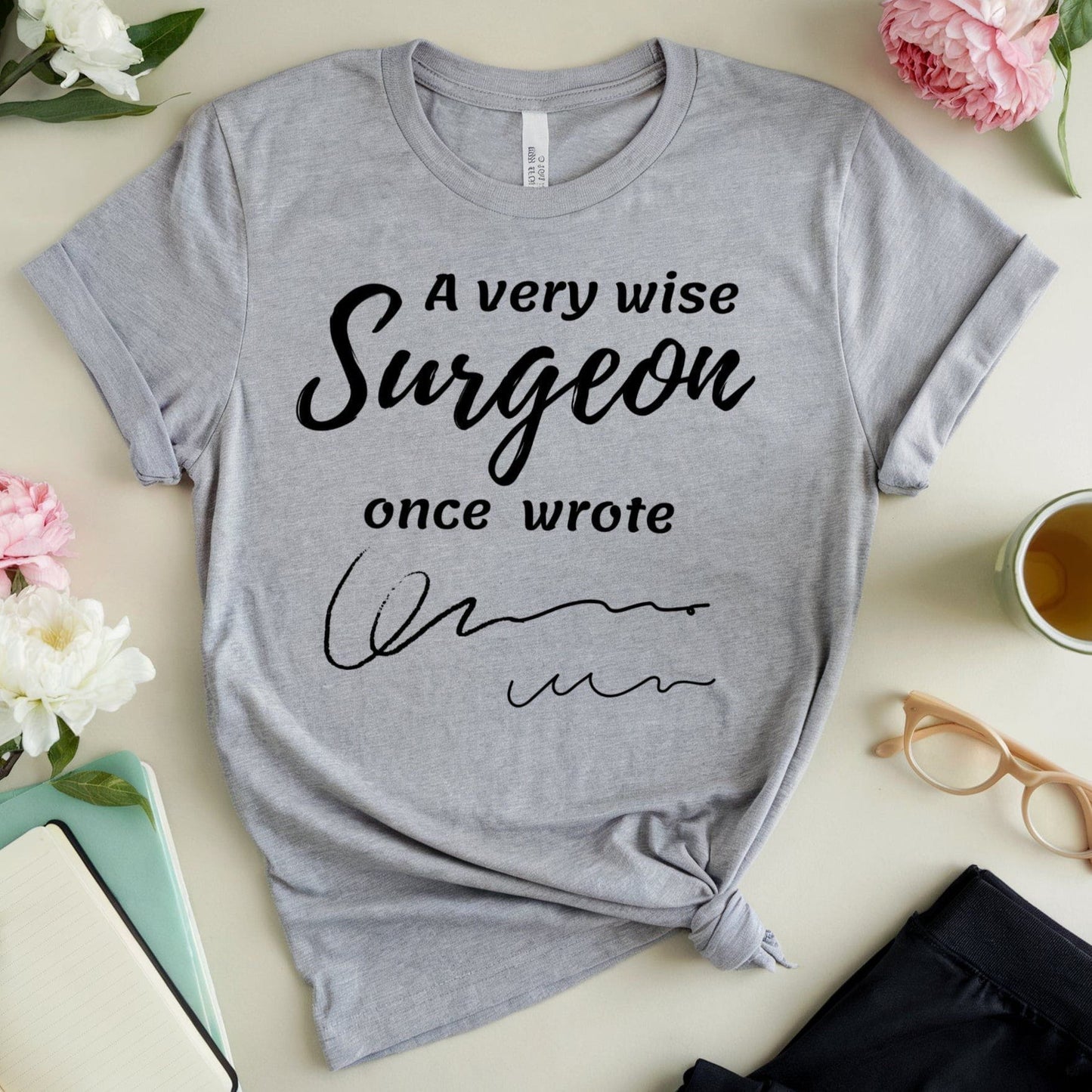 Gray t-shirt  for surgeons with humorous text on a table with flowers and a cup.