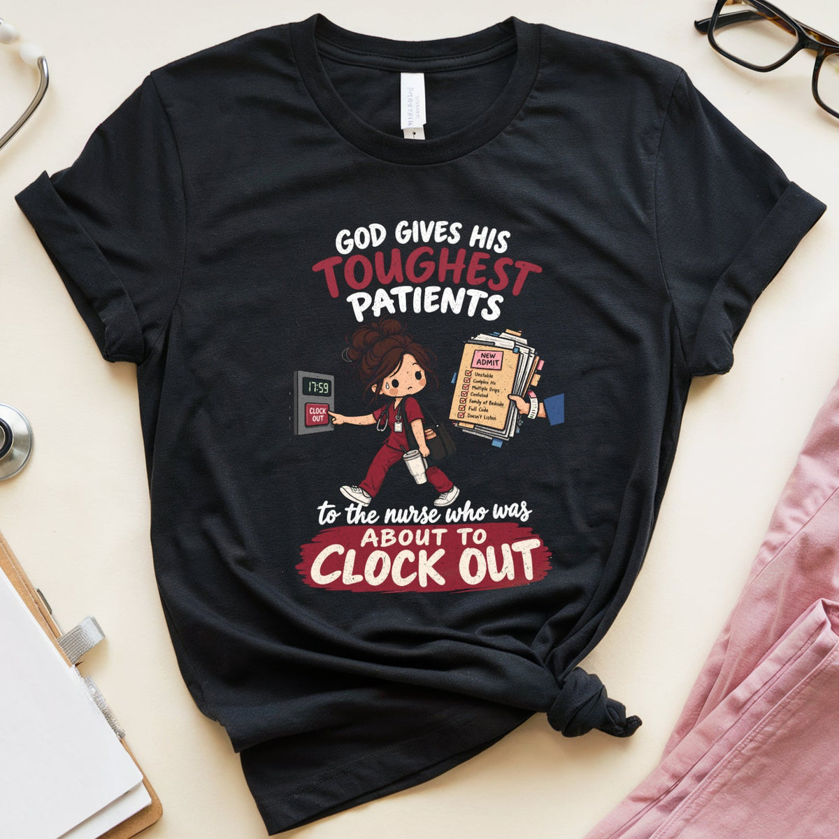 A black graphic t-shirt featuring a cartoon nurse and the text 'GOD GIVES HIS TOUGHEST PATIENTS to the nurse who was ABOUT TO CLOCK OUT', styled in a flat lay with pink scrubs, glasses, a stethoscope, a notebook, and a pen on a light beige background.