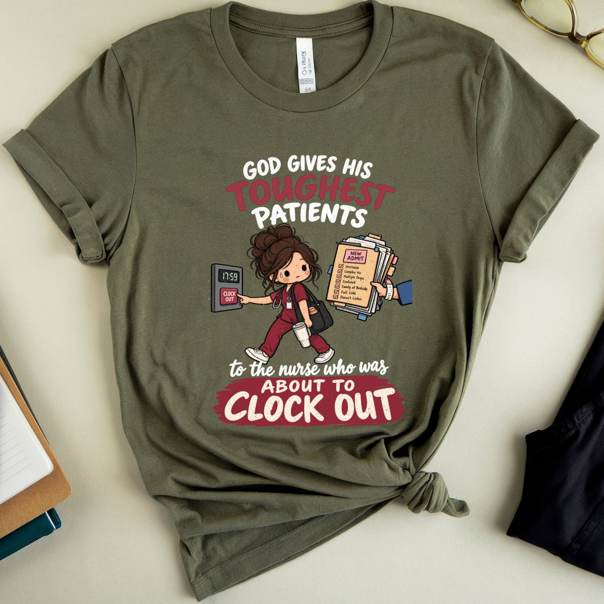 An olive green graphic t-shirt featuring a cartoon nurse and the text 'GOD GIVES HIS TONIGHTY PATIENTS to the nurse who was ABOUT TO CLOCK OUT', styled in a flat lay with glasses, a notebook, and a pair of pants on a light beige background.