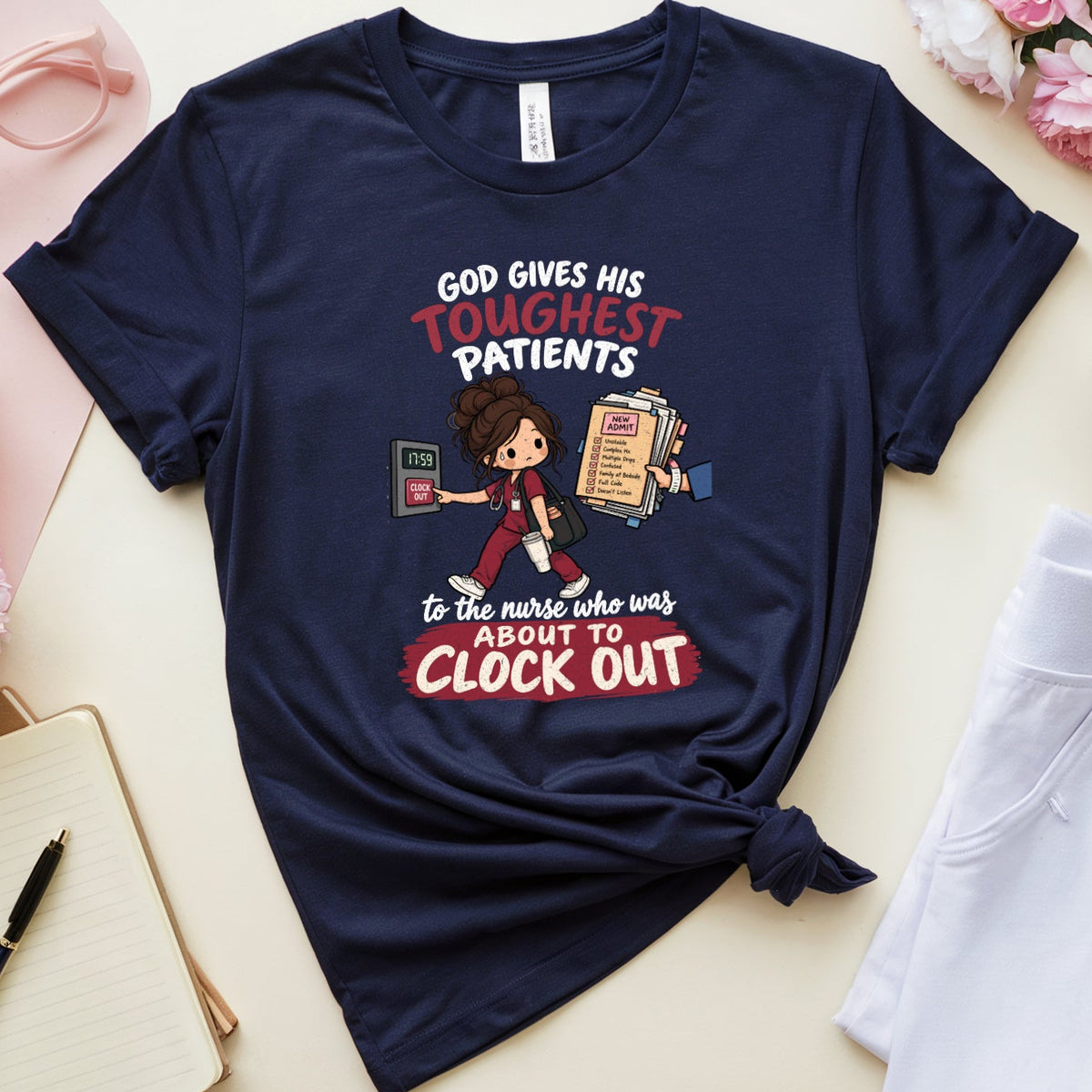 A navy blue graphic t-shirt with a humorous medical quote and cartoon nurse illustration, styled in a flat lay with white pants, a notebook, pink flowers, and a cup on a beige background.