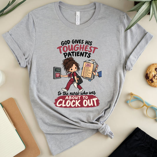A heather gray graphic T-shirt laid flat on a light beige surface, styled with a notebook, pen, glass of milk, cookie, eyeglasses, and plant leaves around it. The shirt has a printed nurse-themed slogan and cartoon nurse graphic.