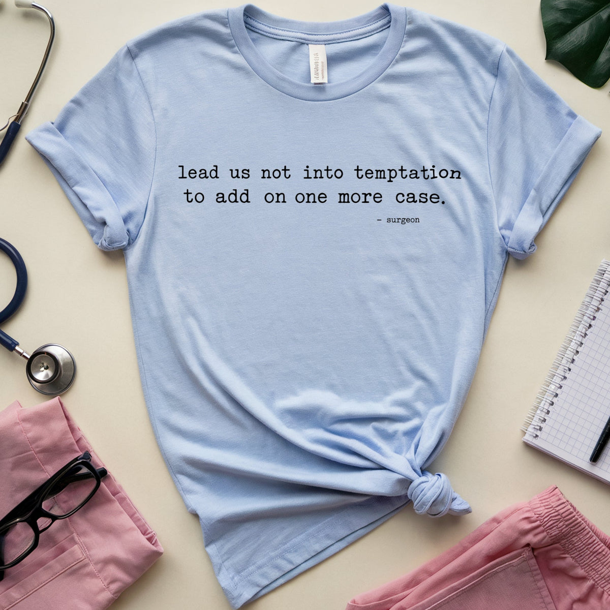 A light blue graphic t-shirt with a black printed quote, styled in a flat lay with medical-themed props including pink scrubs, a stethoscope, glasses, a notebook, and a plant on a beige background.