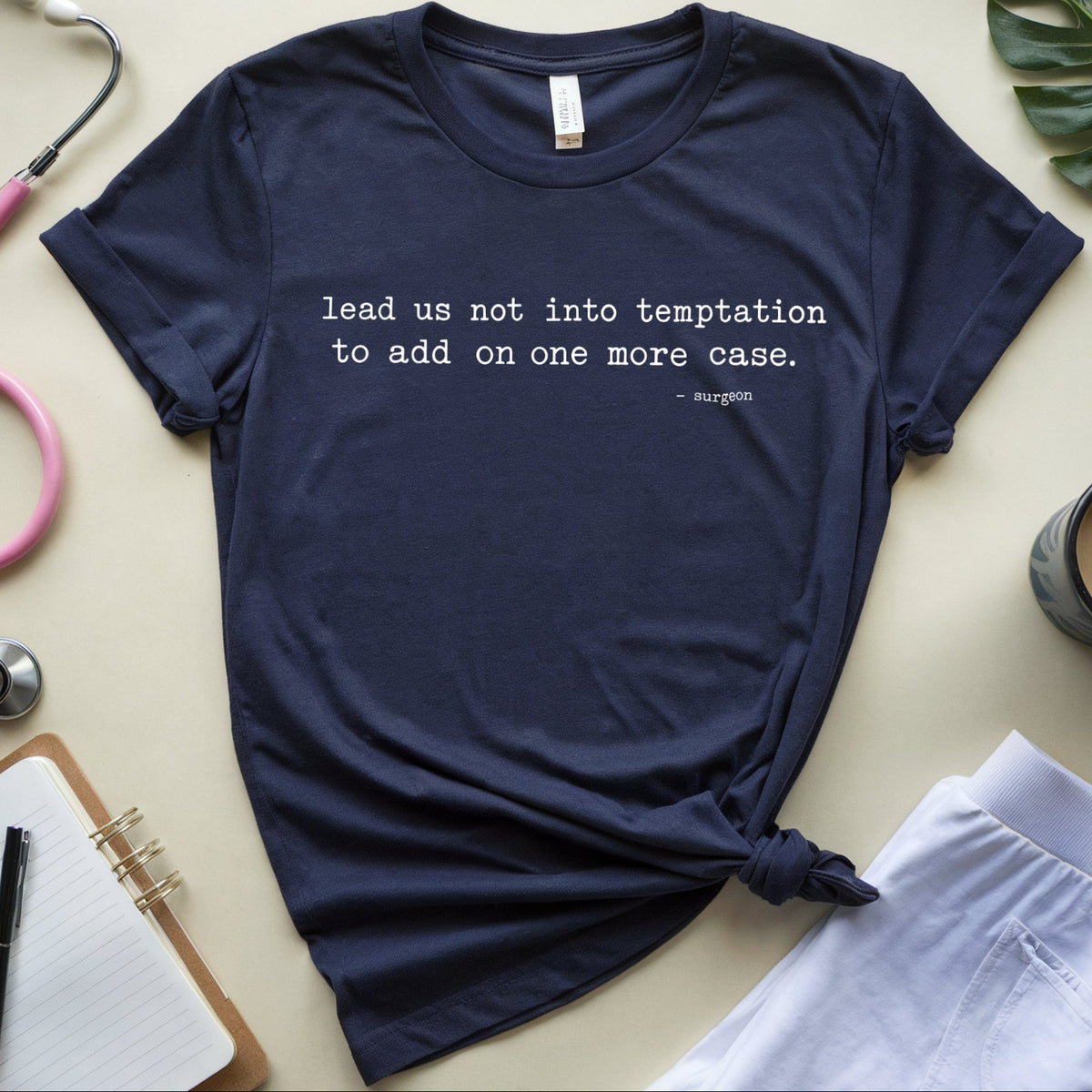 A navy blue graphic t-shirt with a white printed quote by Hippocrates, styled in a flat lay with a pink stethoscope, notebook, coffee cup, and green leaves on a beige background.