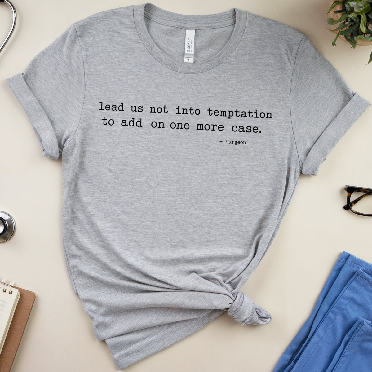 A heather gray short-sleeve T-shirt with rolled sleeves and a knotted hem, laid flat on a light beige surface. The shirt has black printed text reading 'lead us not into temptation to add on one more case.