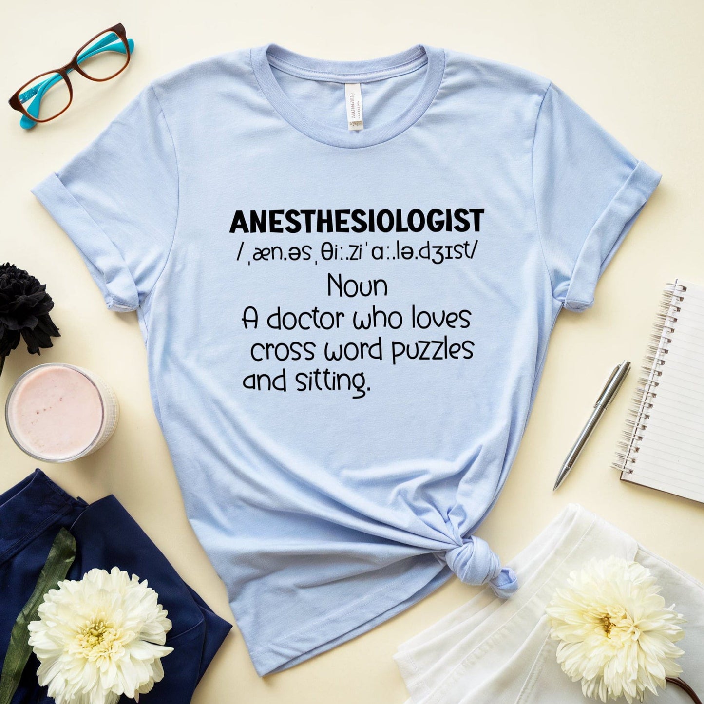 Light blue t-shirt with text about anesthesiologists on a table with flowers, glasses, and a notebook.