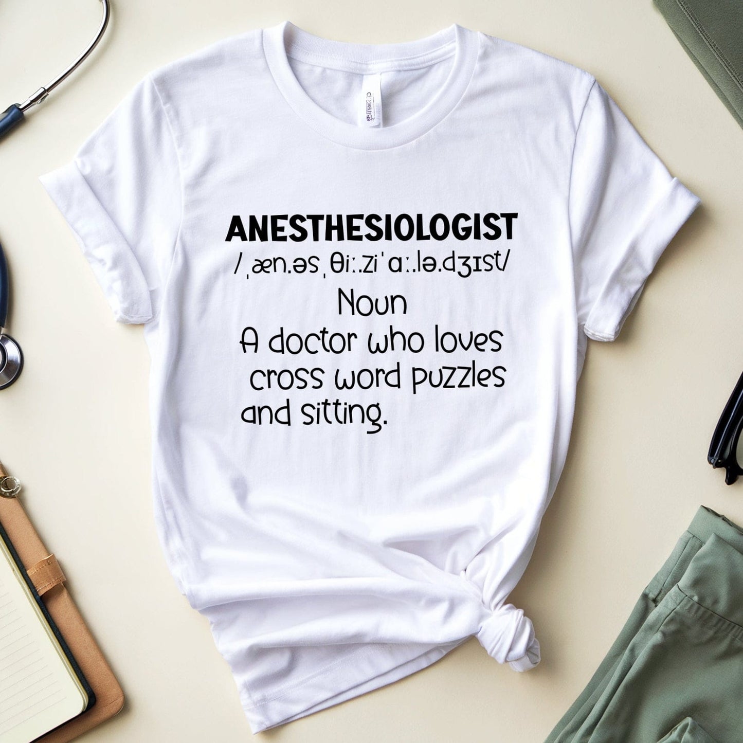 White t-shirt with 'ANESTHESIOLOGIST' definition text on a light surface with medical-themed items.