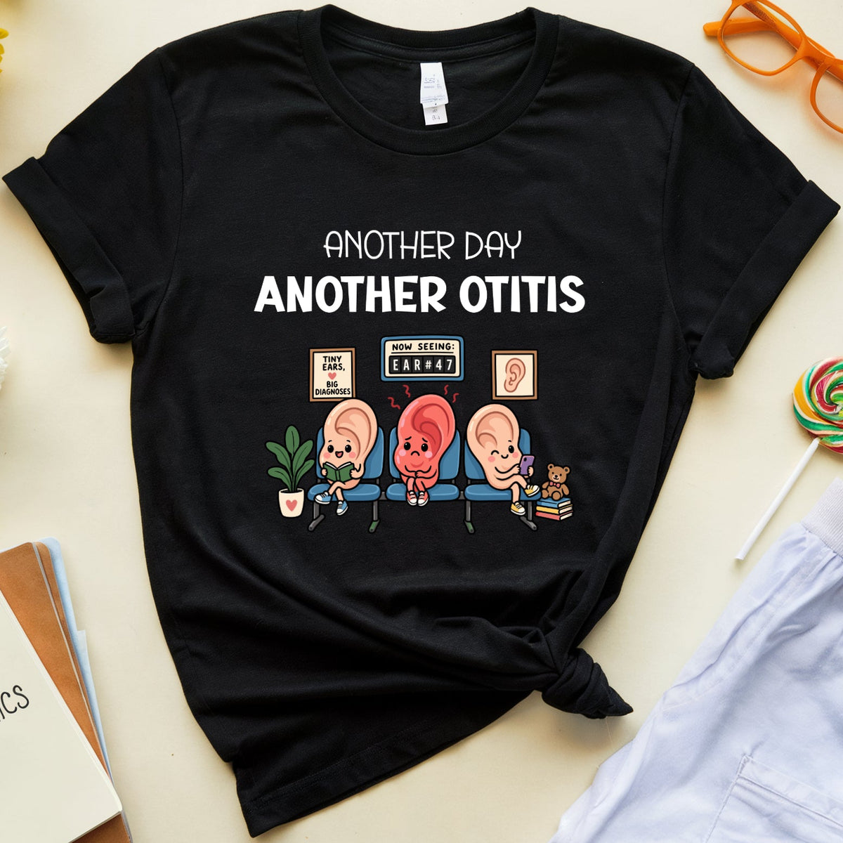 A black graphic t-shirt featuring the text 'ANOTHER DAY ANOTHER OTIS' above three cartoon otitis ears, styled in a flat lay with light blue scrubs, orange glasses, a lollipop, a notebook, and flowers on a beige background.