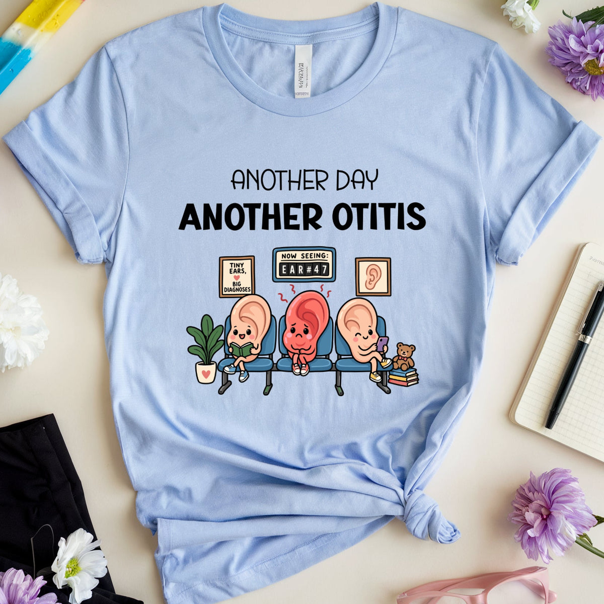 A light blue graphic t-shirt with the text 'ANOTHER DAY ANOTHER OTITIS' and a cartoon illustration of three babies with otitis ears, styled in a flat lay with flowers, a popsicle, glasses, and a notebook.