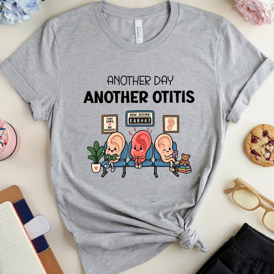 A heather gray graphic t-shirt laid flat on a light beige surface, styled with a knot at the bottom hem. The shirt features a cartoon graphic of three patients sitting on examination chairs with the text 'ANOTHER DAY ANOTHER OTITIS'.