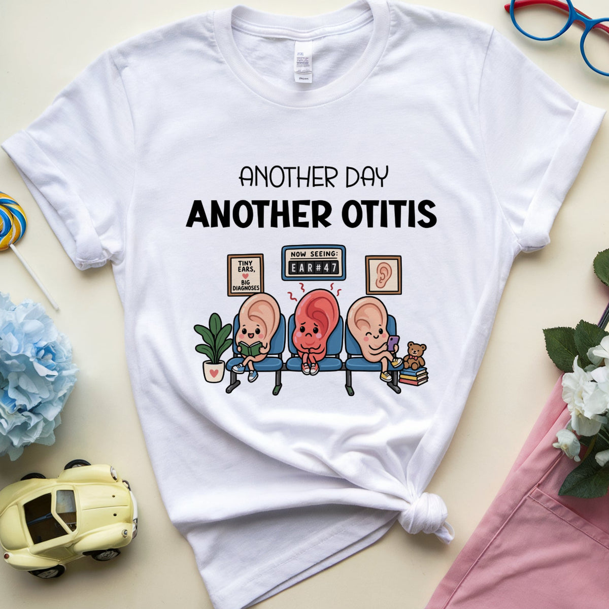 A white graphic t-shirt featuring a cartoon illustration of three patients with otitis symptoms sitting on a exam table, with the text 'ANOTHER DAY ANOTHER OTITIS'. The t-shirt is styled in a flat-lay arrangement with pink pants, blue flowers, red-framed glasses, and a small toy car on a light beige background.