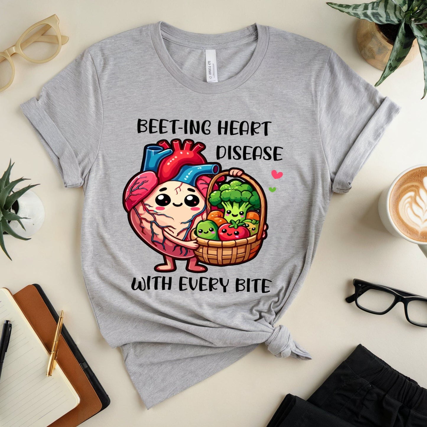 Gray t-shirt with a heart and vegetables graphic and text on a table with a cup of coffee and glasses.
