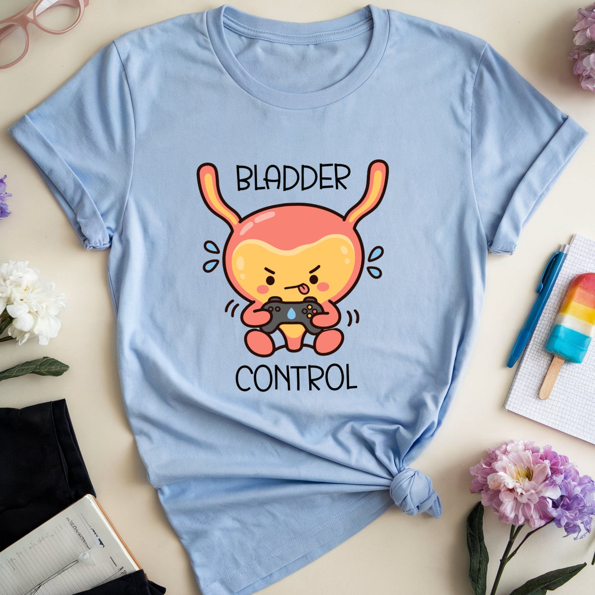 A light blue graphic t-shirt with a cartoon character and the text 'BLADDER CONTROL' is styled in a flat lay on a beige background with flowers, glasses, a notebook, and a popsicle.