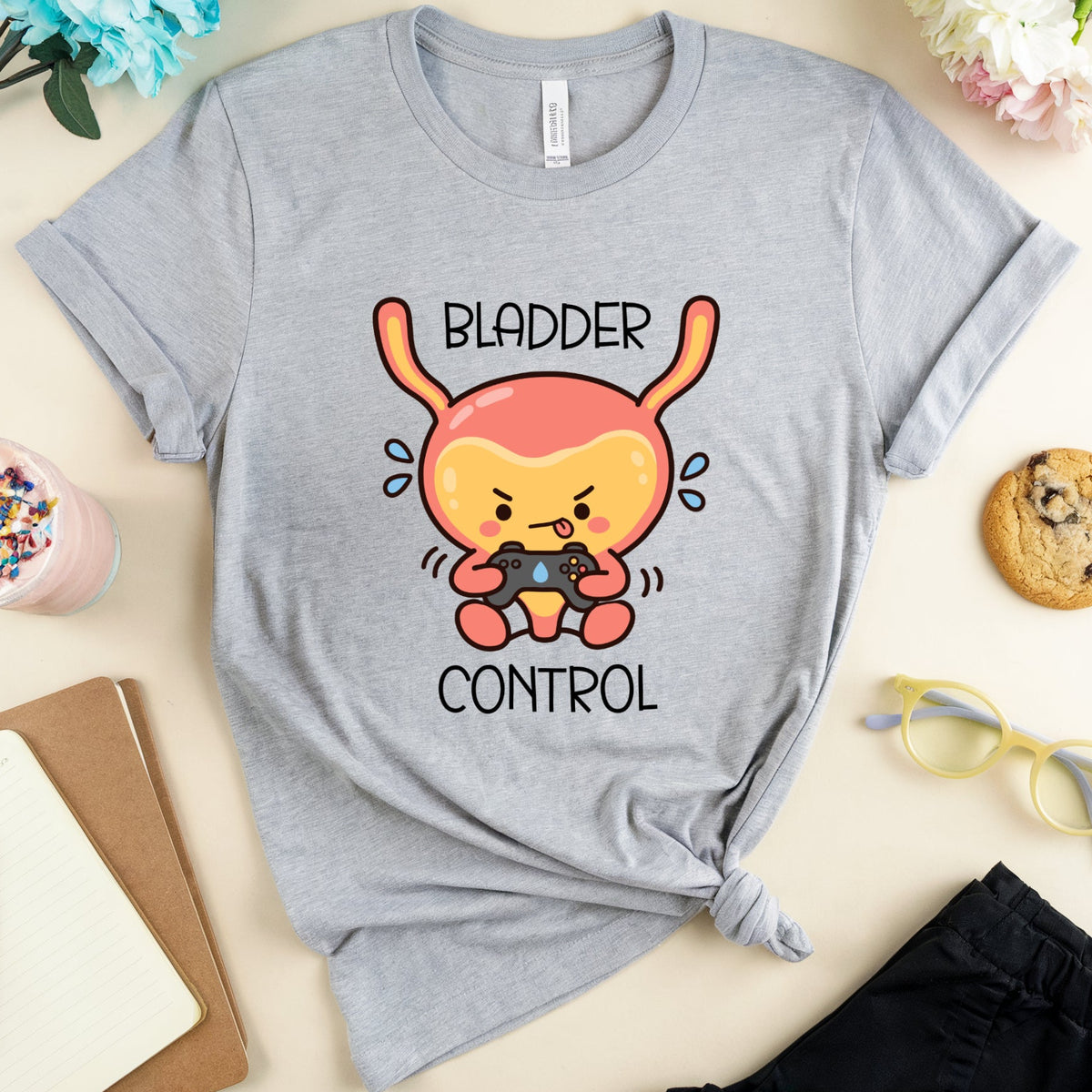 A heather gray graphic t-shirt featuring a cartoon bunny holding a game controller with the words 'BLADDER CONTROL' printed on the front. The t-shirt is styled in a flat lay with a drink, notebook, cookie, glasses, and flowers on a light beige background.