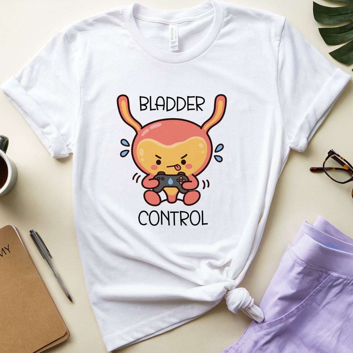 A white graphic t-shirt featuring a cartoon bladder character holding a game controller with the text 'BLADDER CONTROL' printed on the front. The t-shirt is styled in a flat-lay arrangement with a purple pair of shorts, a notebook, eyeglasses, a coffee cup, and a green leaf on a light beige background.