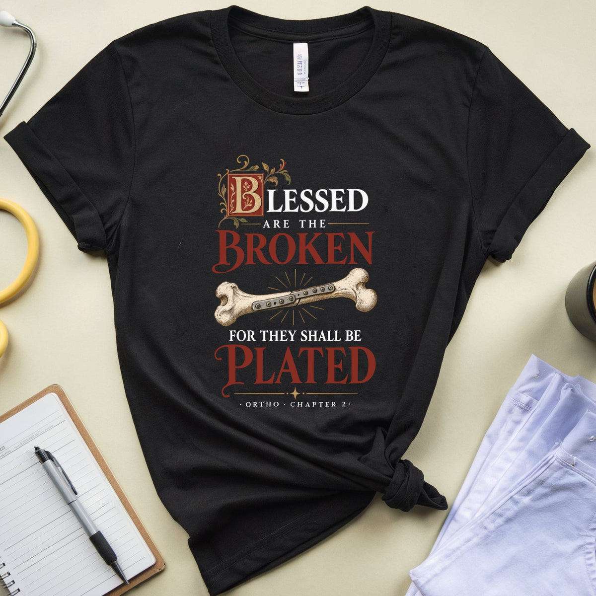 A black graphic t-shirt with a medical-themed quote and bone illustration, styled in a flat lay with a stethoscope, notebook, pen, coffee mug, and folded light blue jeans on a beige background.