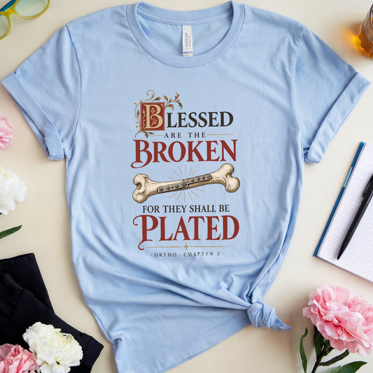 A light blue graphic t-shirt with a distressed print reading 'Blessed are the Broken for they shall be Plated' with a bone illustration, styled in a flat lay with flowers, glasses, a notebook, and a drink on a beige background.