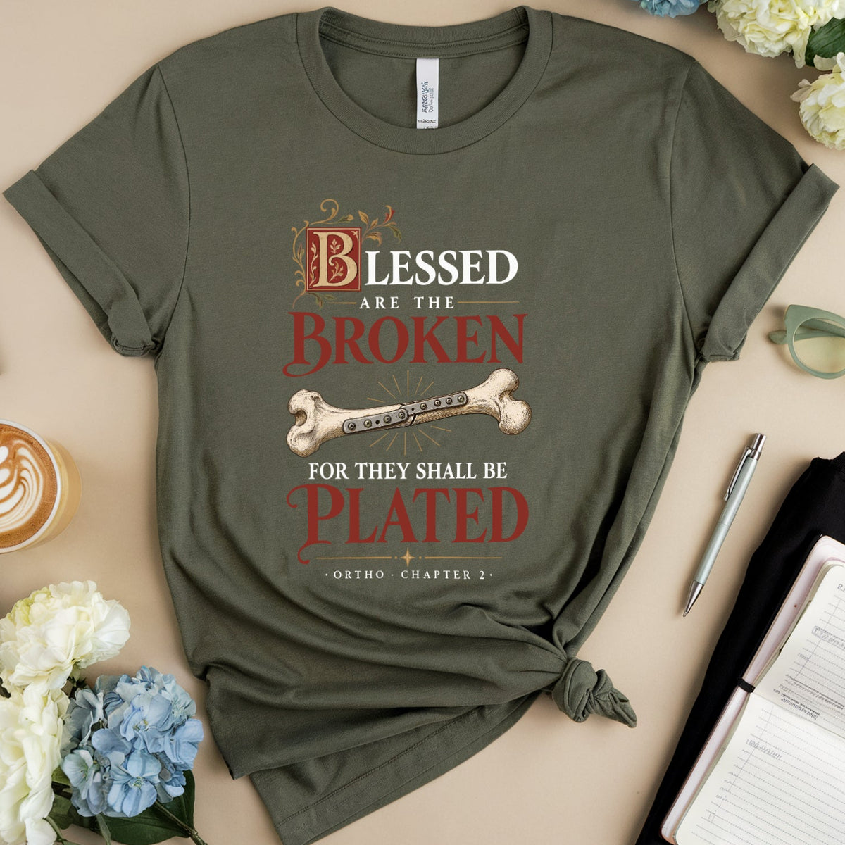 An olive green graphic t-shirt is presented in a styled flat-lay arrangement on a beige background. The shirt features a printed design with the text 'Blessed are the Broken for they shall be Plated' and a bone graphic.