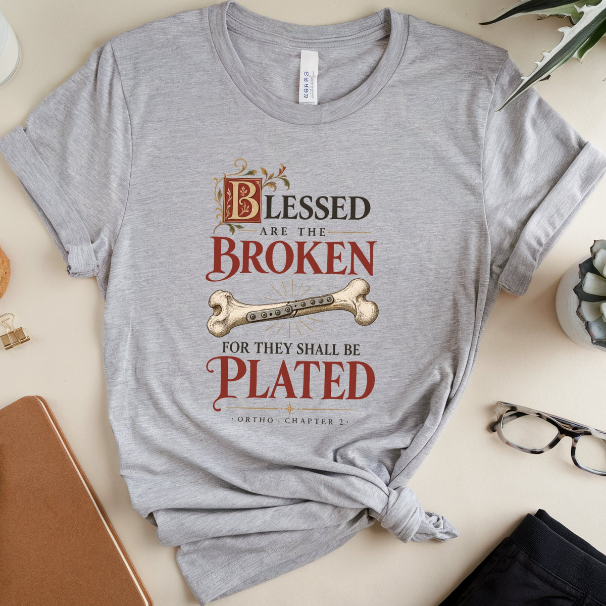 A heather gray graphic t-shirt with a knotted hem, featuring the text 'Blessed are the Broken for they shall be Plated' with a bone graphic. The t-shirt is styled in a flat-lay arrangement on a beige background with a notebook, glasses, a cookie, and plants.