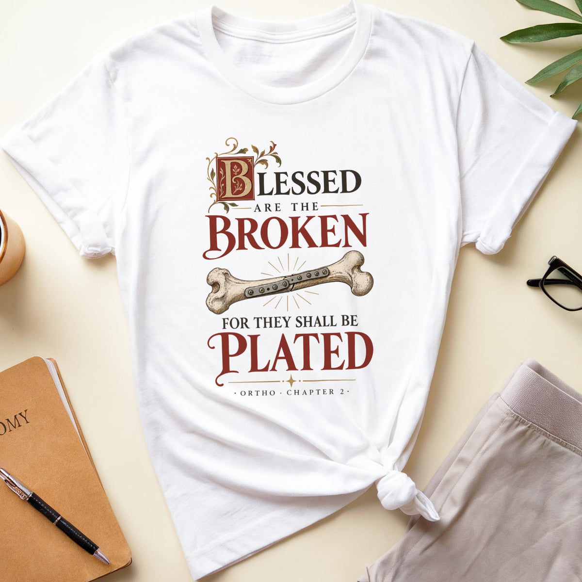A white graphic t-shirt with a humorous orthopedic quote printed on the front, styled in a flat lay with a notebook, coffee mug, eyeglasses, and shorts on a light beige background.
