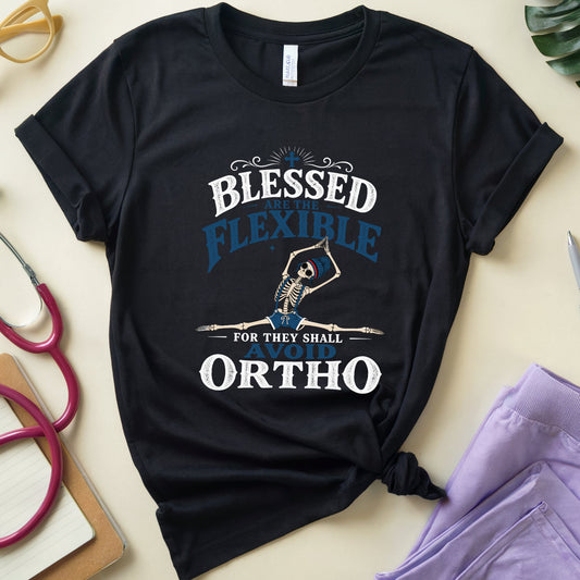 A black graphic t-shirt with a medical-themed print reading 'BLESSED FLEXIBLE FOR THEY SHALL ORTHO' with a skeleton illustration, styled in a flat-lay arrangement with a stethoscope, glasses, notebook, and purple pants on a beige background.