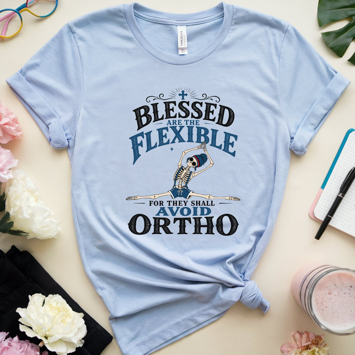 A light blue graphic t-shirt with a knotted hem, featuring a skeleton flexing with the text 'BLESSED AS THE FLEXIBLE FOR THEY SHALL AVOID ORTHO'. The t-shirt is styled in a flat-lay arrangement with pink and white flowers, a pair of colorful sunglasses, a black notebook, and a drink on a light beige background.