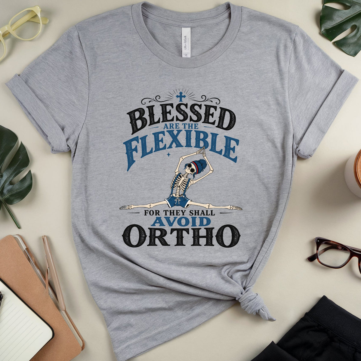 A heather gray graphic t-shirt with a 'Blessed Are The Flexible For They Shall Avoid Ortho' skeleton dancing slogan, styled in a flat lay with glasses, a coffee cup, notebook, and leaves on a beige background.