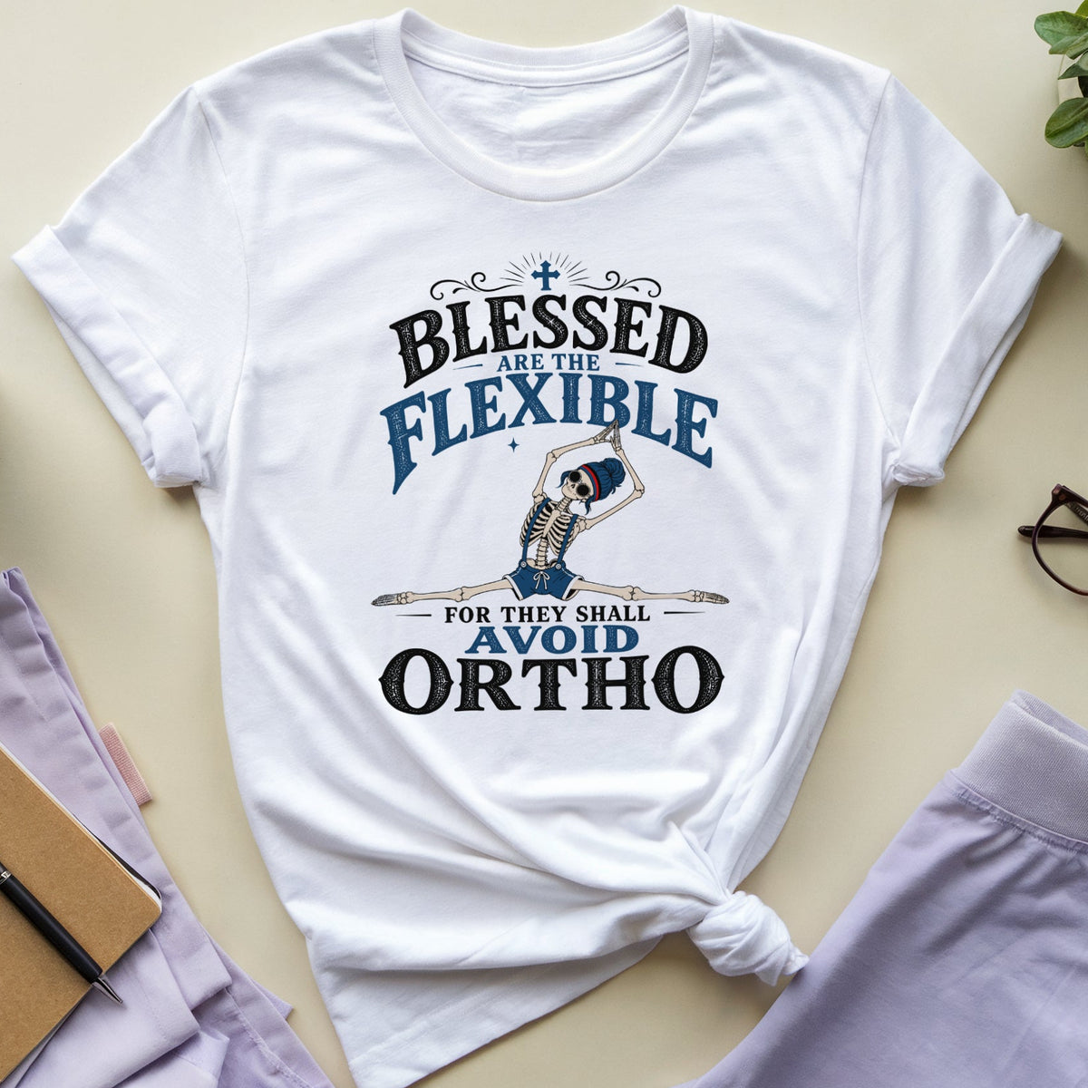 A white graphic t-shirt with rolled sleeves, featuring a blue and black print that reads 'BLESSED ARE THE FLEXIBLE FOR THEY SHALL AVOID ORTHO' alongside an illustration of a gymnast. The shirt is styled in a flat-lay arrangement on a beige background with lavender pants, glasses, a notebook, a plant, and a coffee mug.