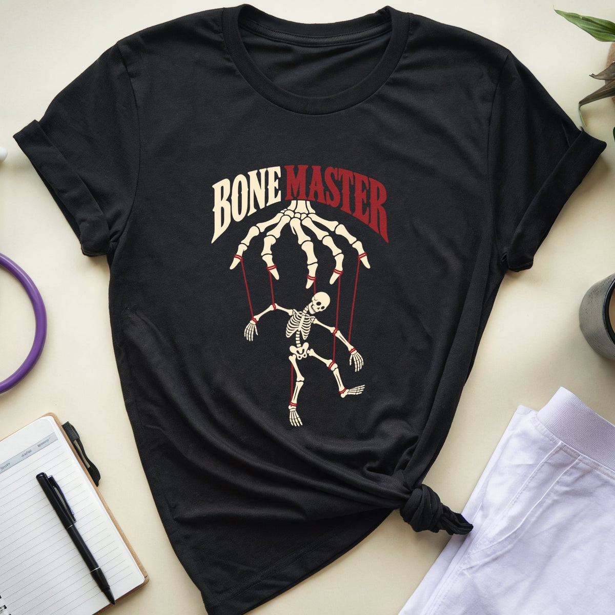 A black graphic t-shirt featuring a marionette skeleton design with the text 'BONE MASTER' in cream and red, styled in a flat lay with medical-themed props like a stethoscope, notebook, and coffee cup on a beige background.