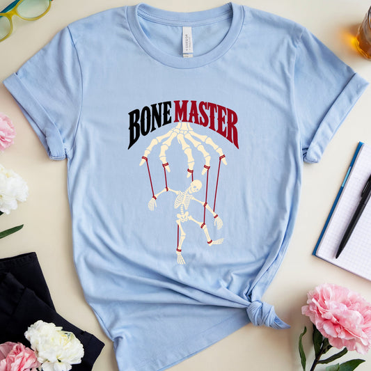 A light blue graphic t-shirt with 'BONE MASTER' and a skeleton hand design, styled in a flat lay with flowers, glasses, a notebook, and a drink on a beige background.