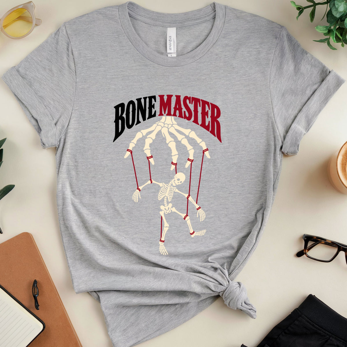 A heather gray graphic t-shirt featuring a skeleton hand gripping maracas under the text 'BONE MASTER' in black and red. The t-shirt is styled in a flat lay with a knot at the bottom, surrounded by lifestyle props including sunglasses, a notebook, glasses, and a coffee cup on a light beige background.