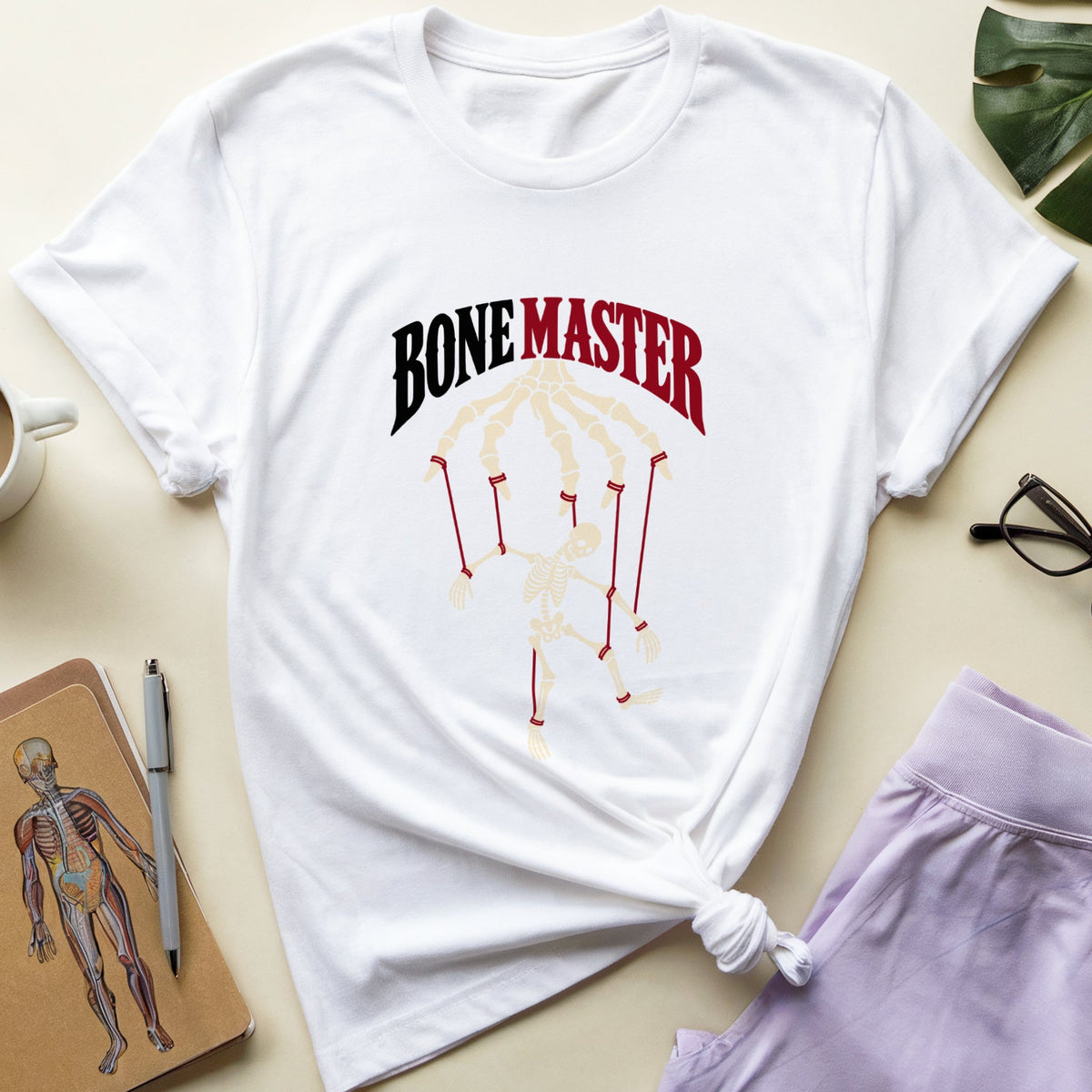 A white graphic t-shirt with 'BONE MASTER' printed in black and red above a beige skeleton hand design, styled in a flat lay with a cup of coffee, anatomical notebook, glasses, and lavender shorts on a light beige background.