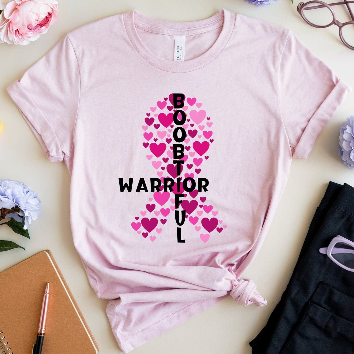 Pink t-shirt with a pink ribbon design and text on a light surface with flowers and glasses.