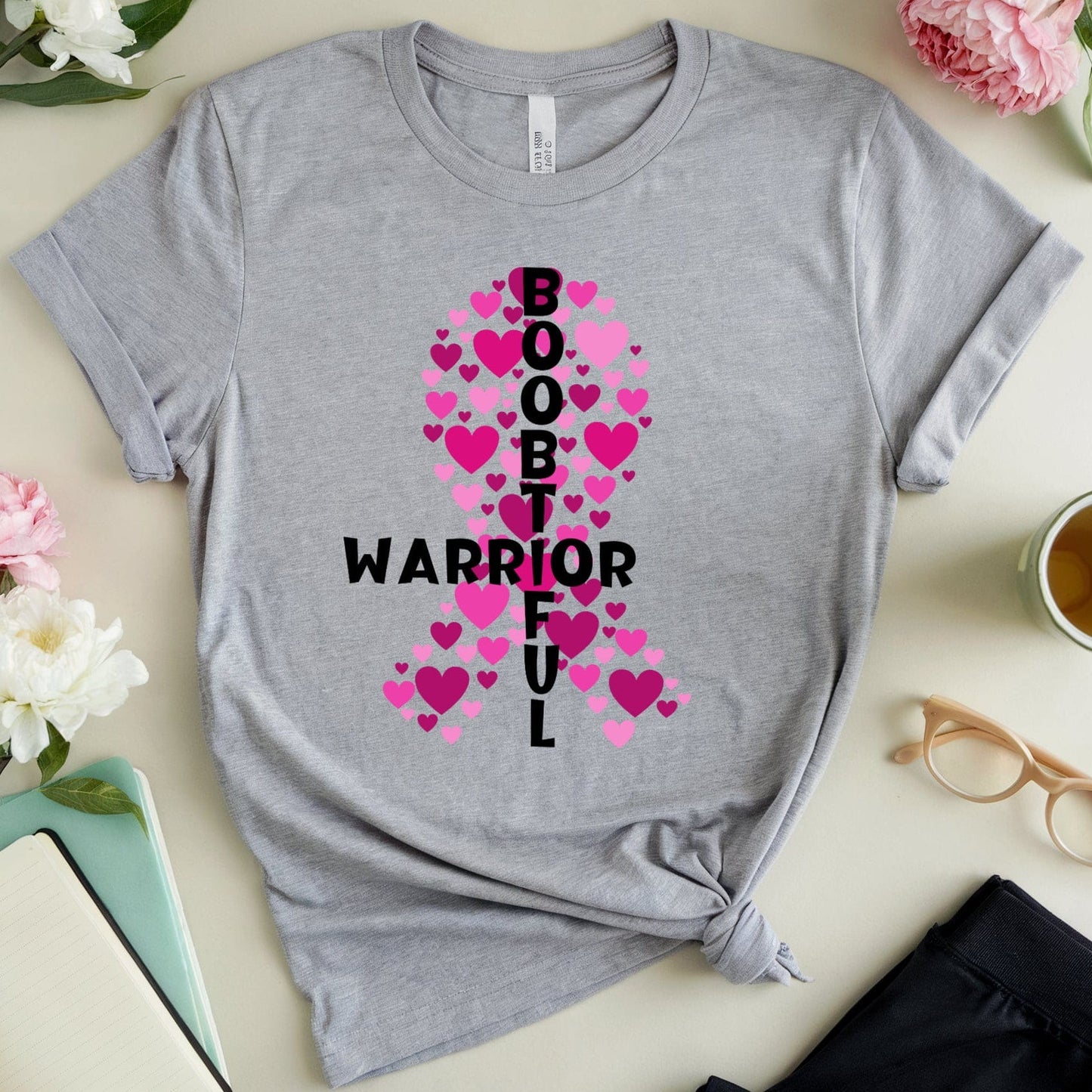 Gray t-shirt with pink ribbon and text design on a light surface with flowers and a cup.