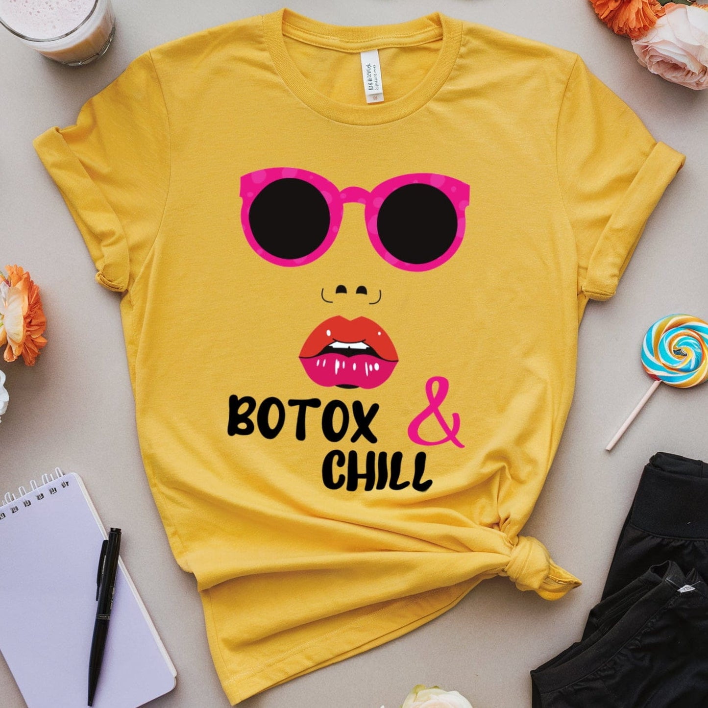 Yellow t-shirt with sunglasses and lips graphic, surrounded by flowers and a drink on a light surface.