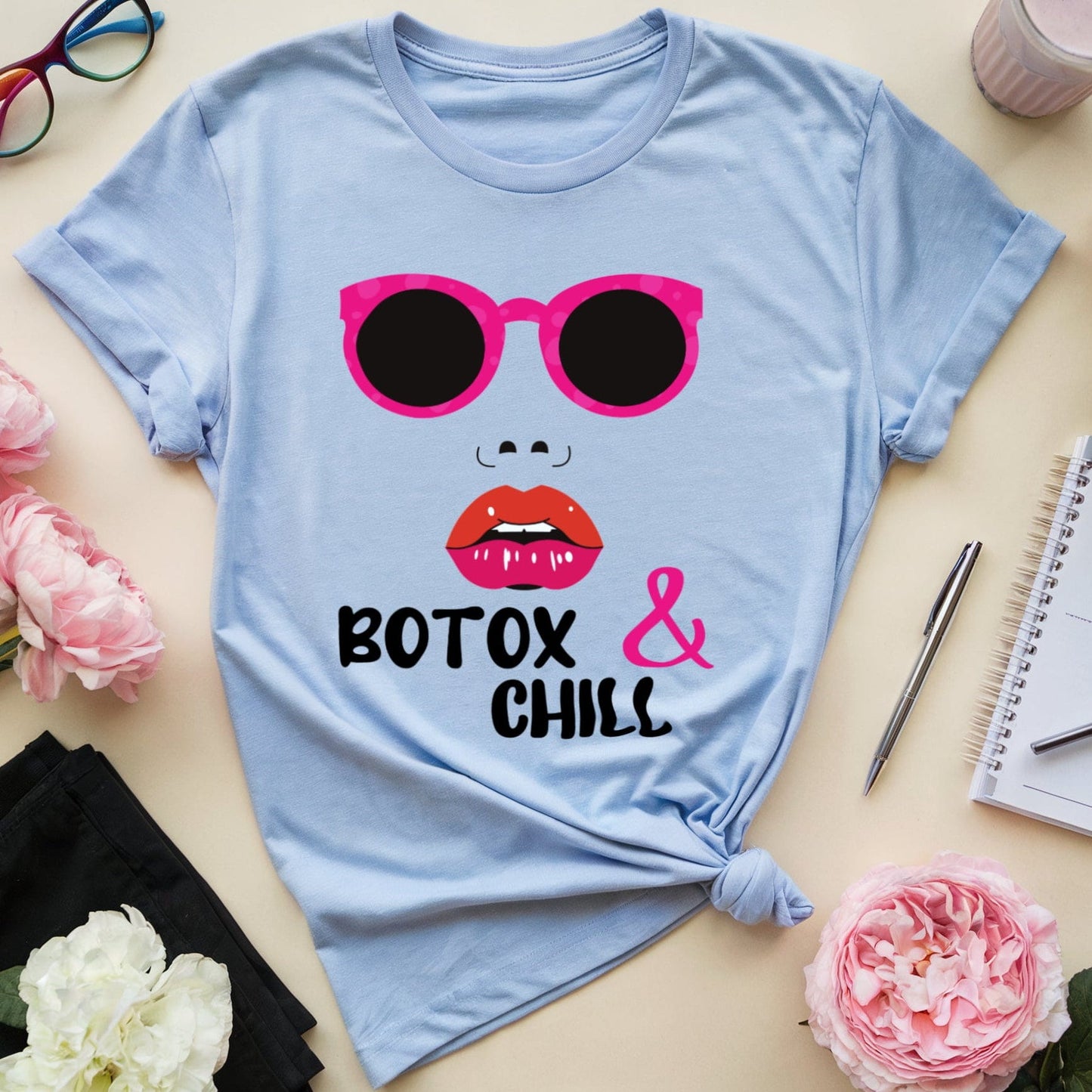 Light blue t-shirt with sunglasses and lips graphic and 'Botox & Chill' text on a table with flowers and glasses.