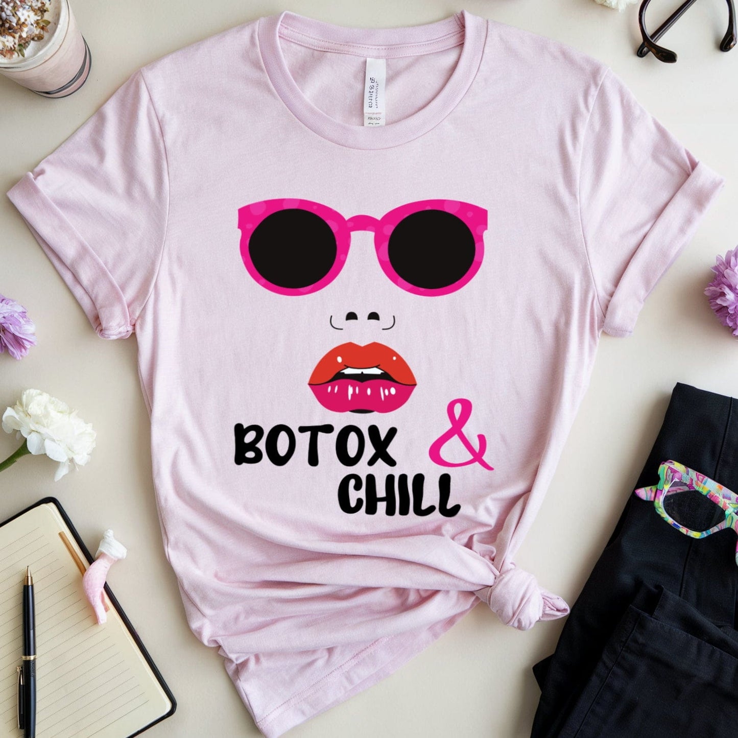 Pink t-shirt with sunglasses and lips graphic and text 'Botox & Chill' on a light surface with flowers and accessories.