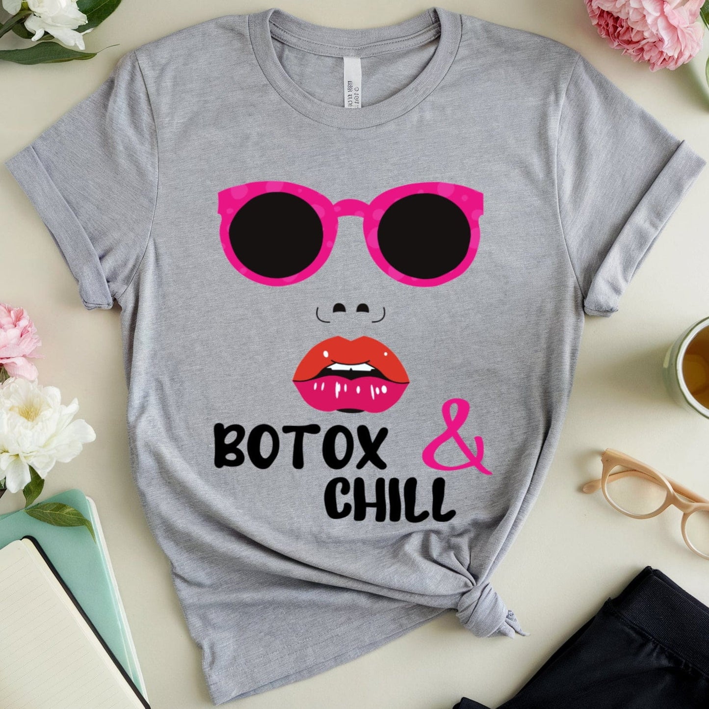 Gray t-shirt with pink sunglasses, red lips, and 'Botox & Chill' text on a light surface with flowers and a cup.