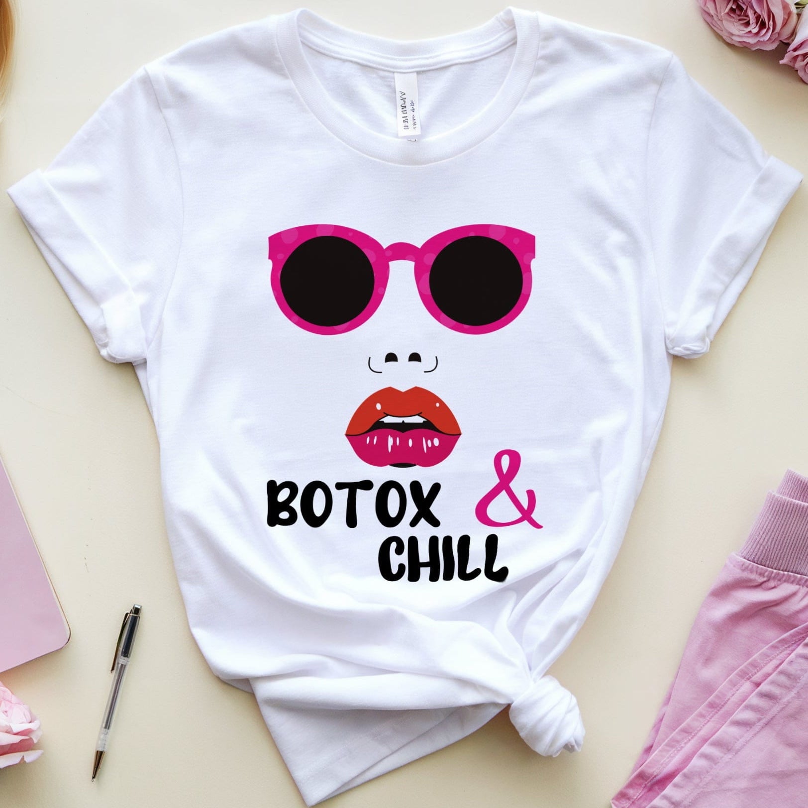 White t-shirt with sunglasses and lips graphic, surrounded by a Barbie doll, notebook, pen, and flowers on a beige surface.