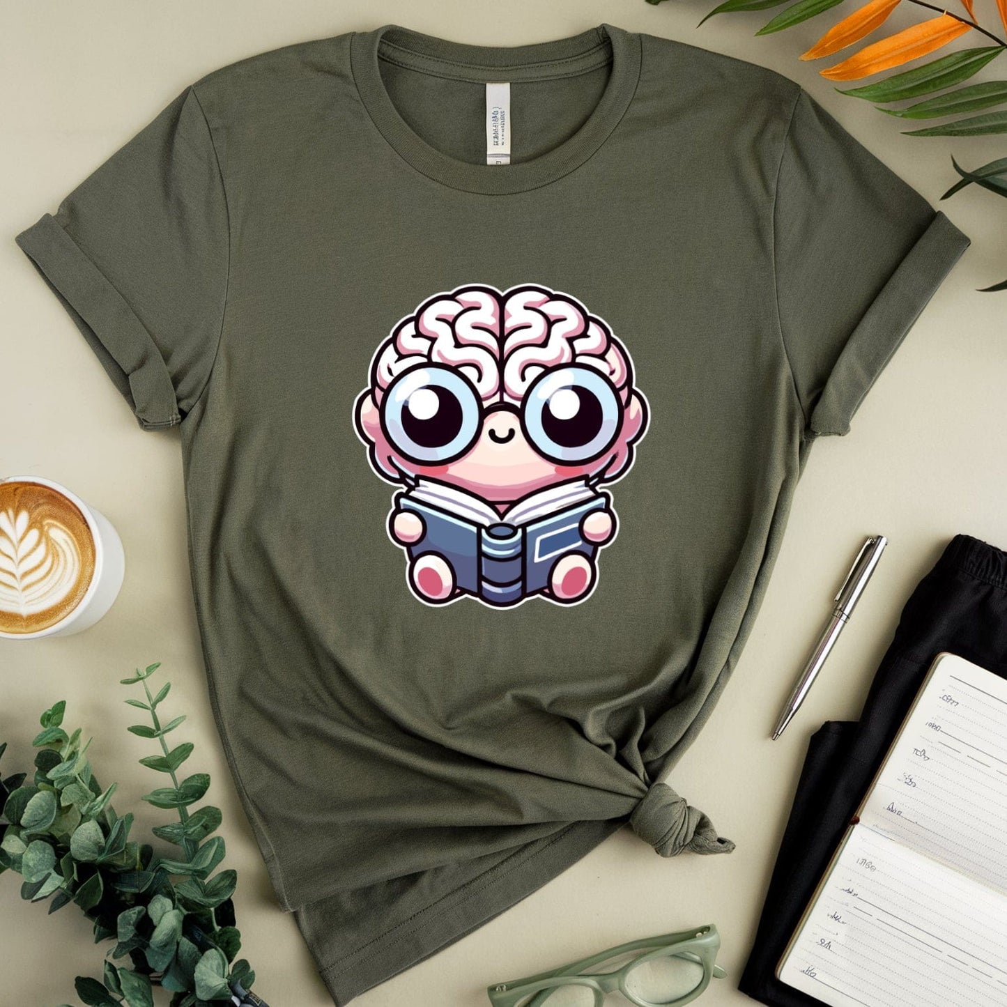 Green t-shirt with a brain character reading a book design, placed on a light surface with a cup of coffee, glasses, and a plant.