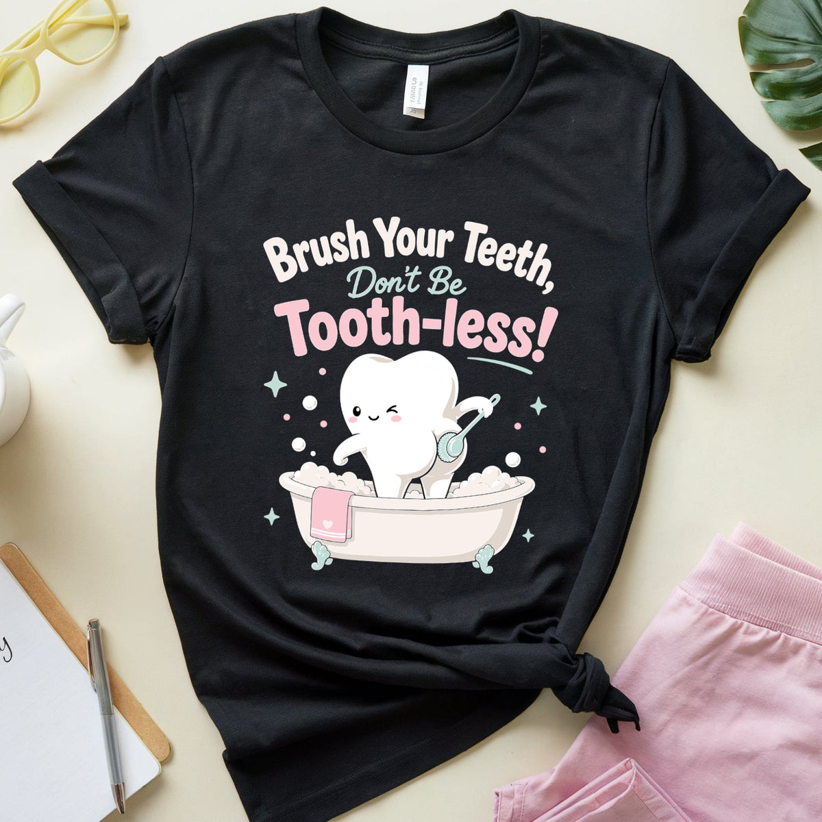 A black graphic t-shirt featuring a cartoon tooth character in a bathtub with the text 'Brush Your Teeth, Don't Be Tooth-less!' in white and pink lettering. The t-shirt is styled in a flat-lay arrangement on a light beige background with pink shorts, yellow sunglasses, a coffee cup, a notebook, and a green leaf.