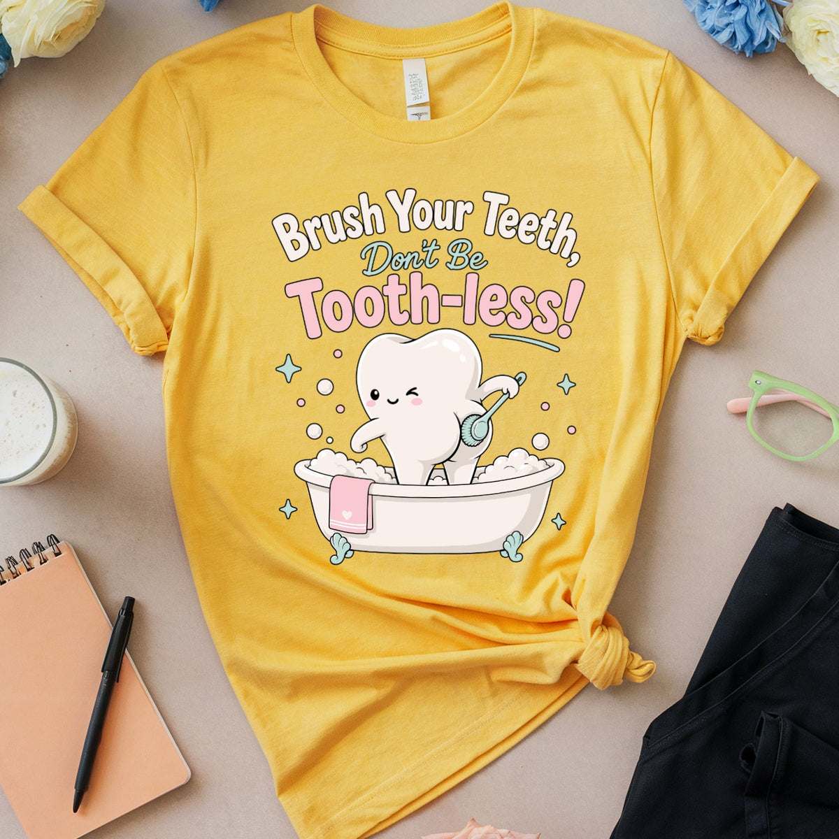 A yellow graphic T-shirt laid flat on a light beige surface, surrounded by flowers, a notebook, glasses, cookies, and a cup. The shirt has a cartoon tooth sitting in a bathtub with the text 'Brush Your Teeth, Don't Be Tooth-less!' printed on the front.