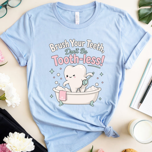 A light blue graphic t-shirt laid flat on a beige surface, surrounded by lifestyle props including pink and white flowers, eyeglasses, a notebook with a pen, a cookie, and a glass of milk. The t-shirt features a cartoon graphic of a tooth character sitting in a bathtub with the text 'Brush Your Teeth, Don't Be Tooth-less!'.