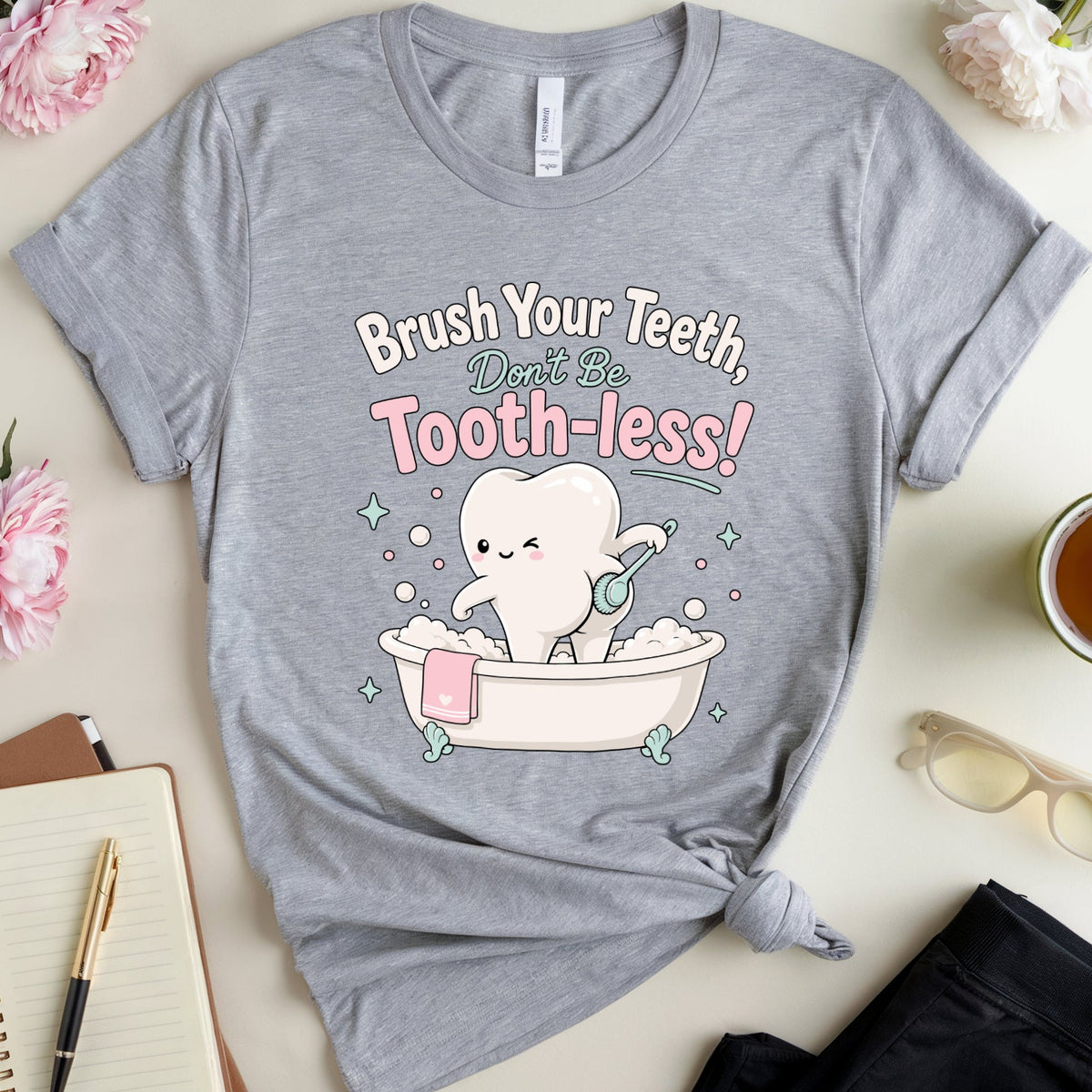 A heather gray graphic T-shirt laid flat on a light beige surface, surrounded by pink flowers, a notebook with a pen, a cup of tea, and folded dark pants. The shirt has a cartoon tooth in a bubble bath with the text 'Brush Your Teeth, Don't Be Tooth-less!' printed on the front.