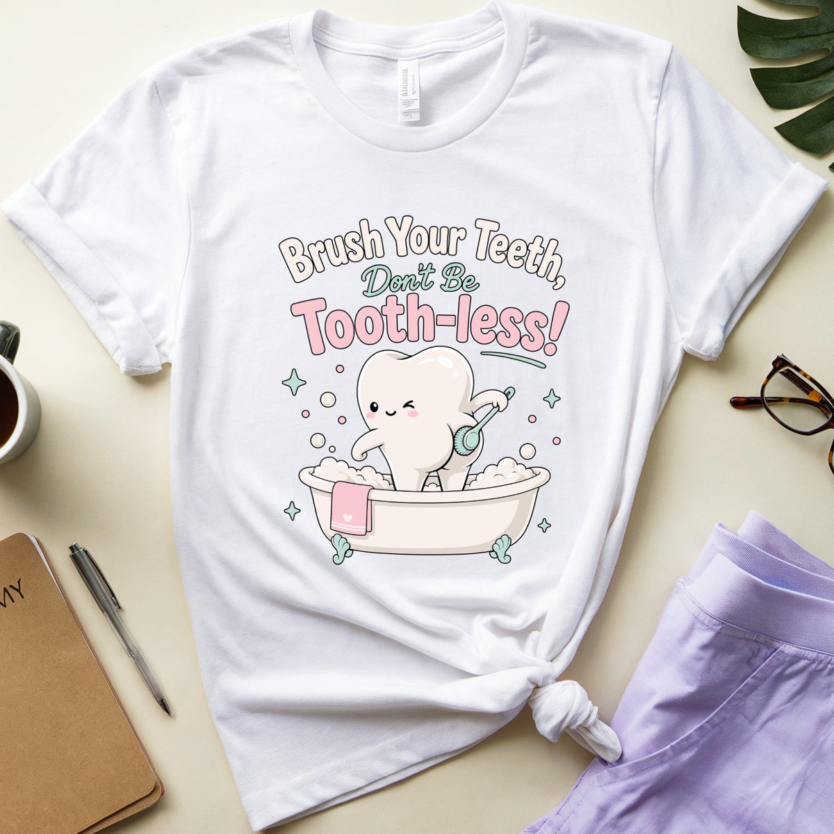 A white graphic t-shirt featuring a cute cartoon tooth character sitting in a bathtub with the text 'Brush Your Teeth, Don't Be Tooth-Jess!' in pink and gray lettering. The t-shirt is styled in a flat-lay arrangement on a light beige background alongside a cup of coffee, a notebook, eyeglasses, and lavender shorts.