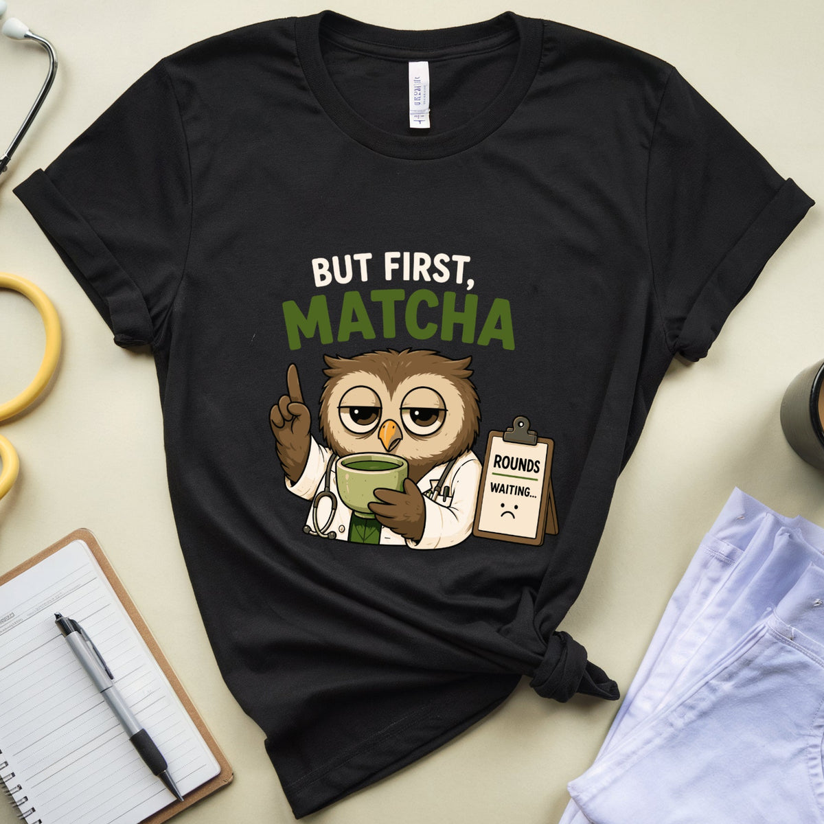 A black graphic t-shirt laid flat on a beige surface, styled with a yellow stethoscope, notebook, pen, folded light blue jeans, and a coffee mug. The shirt features a cartoon owl dressed as a doctor holding a green cup, with the text 'BUT FIRST, MATCHA'.