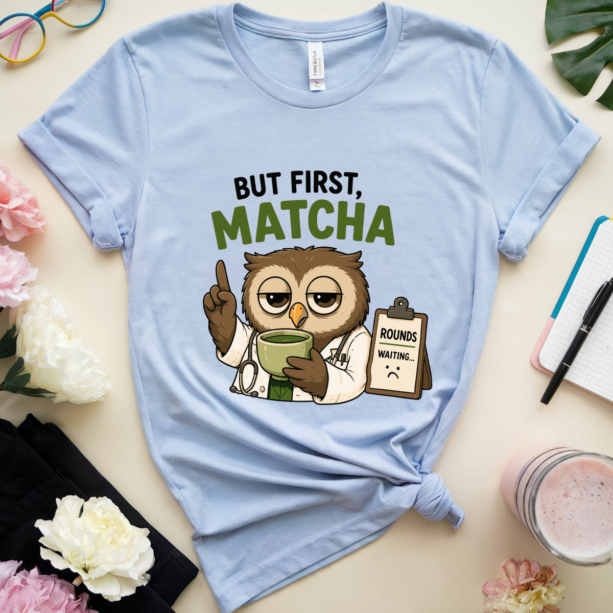 A light blue graphic t-shirt laid flat on a beige surface, styled with flowers, glasses, a notebook, a drink, and a black cloth. The shirt features a cartoon owl doctor holding a green cup with the text 'BUT FIRST, MATCHA'.