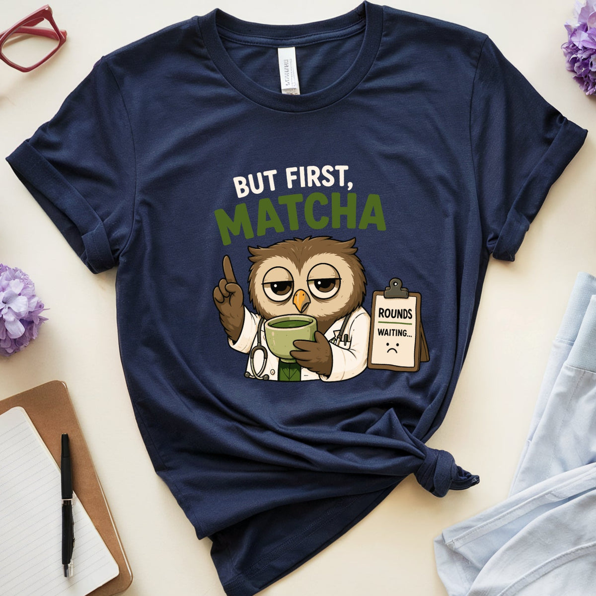 A navy blue graphic t-shirt featuring an owl doctor holding a green cup with the text 'BUT FIRST, MATCHA' in white and green. The t-shirt is styled in a flat lay with light blue pajama pants, a mug of coffee, purple flowers, red eyeglasses, a notebook, and a pen on a light beige background.
