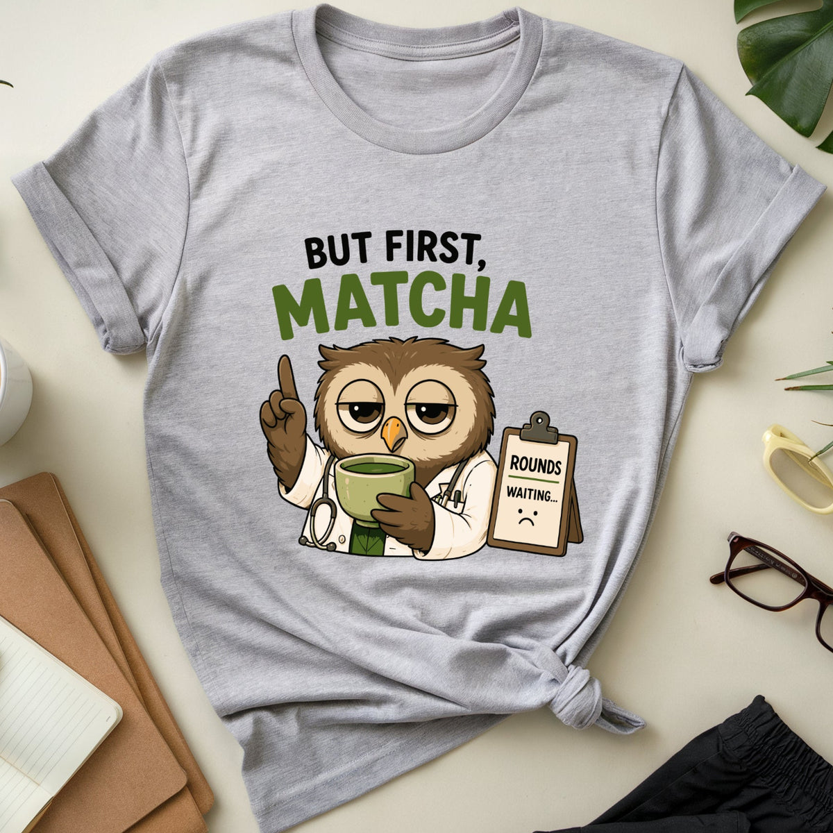 A light heather gray graphic T-shirt laid flat on a cream tabletop, surrounded by lifestyle props including a cup of latte, notebook, plant leaves, sunglasses, glasses, and folded dark pants. The shirt graphic shows a cartoon owl dressed as a doctor holding a mug, with the text 'BUT FIRST, MATCHA' above it.