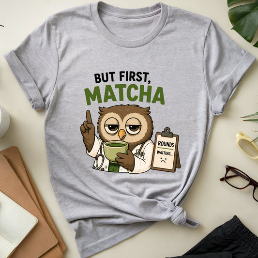 A light heather gray graphic T-shirt laid flat on a cream tabletop, surrounded by lifestyle props including a cup of latte, notebook, plant leaves, sunglasses, glasses, and folded dark pants. The shirt graphic shows a cartoon owl dressed as a doctor holding a mug, with the text 'BUT FIRST, MATCHA' above it.