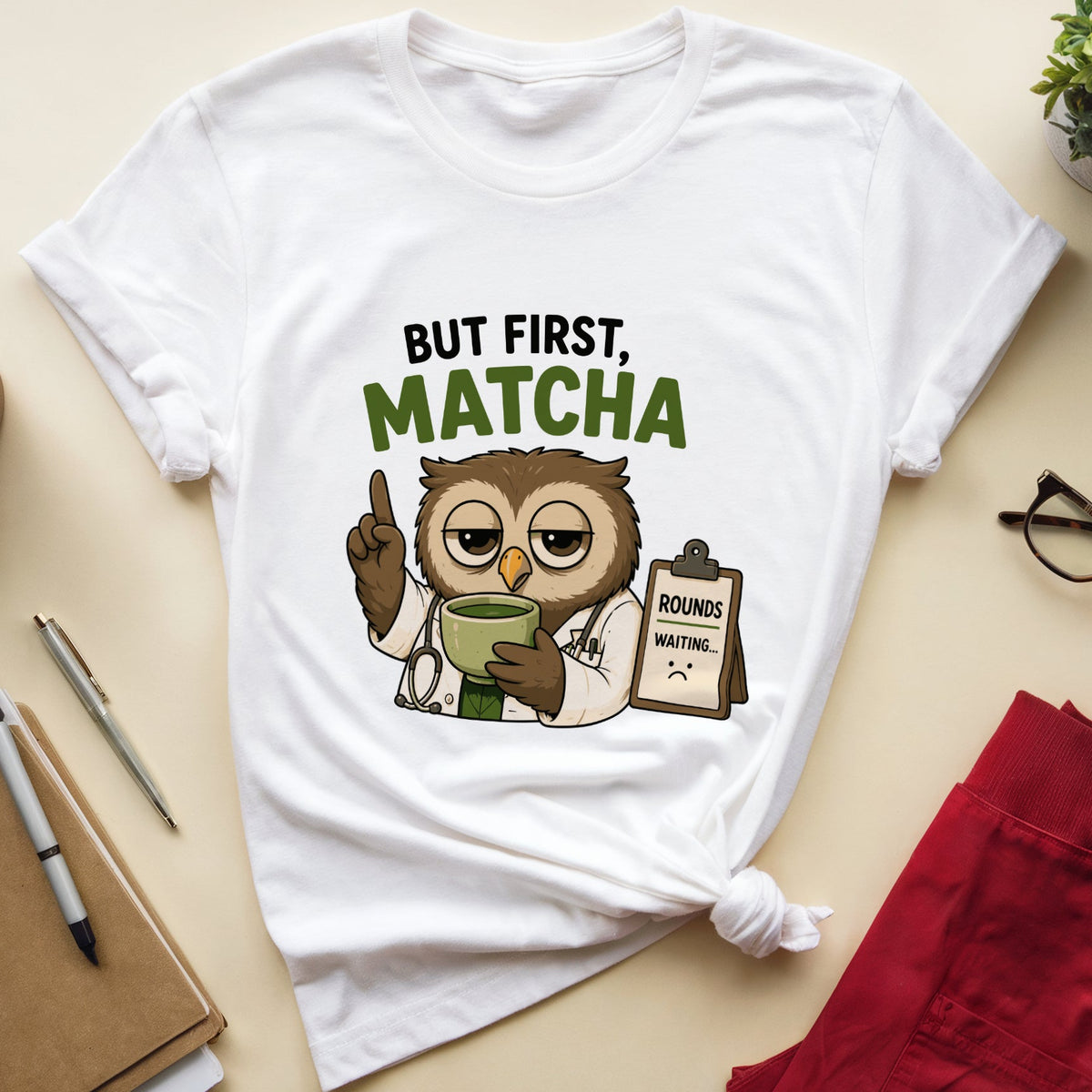 A white graphic t-shirt featuring a cartoon owl dressed as a matcha master with the text 'BUT FIRST, MATCHA', styled in a flat-lay arrangement with red pants, glasses, a notebook, and a small plant on a beige background.